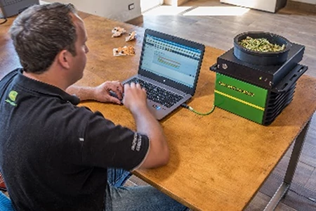A man seated at a table works on a laptop connected to a green and black HarvesLab unit holding a sample of chopped plant material