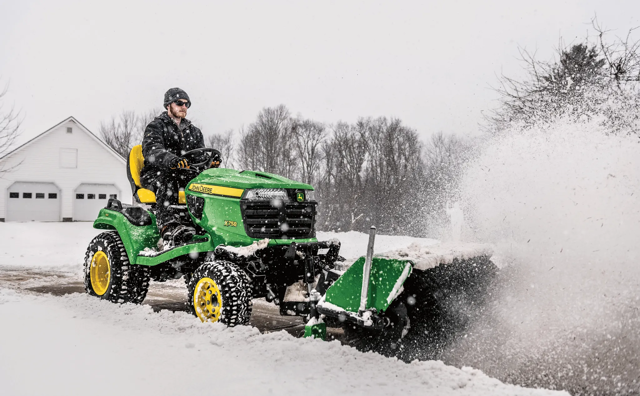 Front right view of an operator on an X758 lawn tractor blowing snow from a driveway with front-attached 52-in. rotary broom