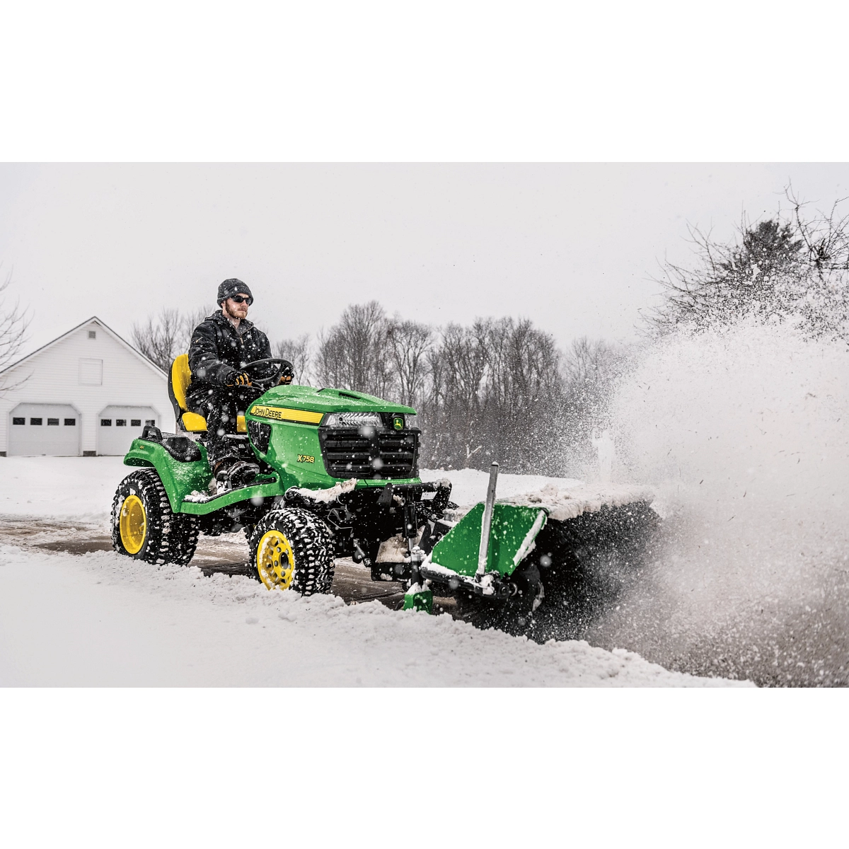 Front right view of an operator on an X758 lawn tractor blowing snow from a driveway with front-attached 52-in. rotary broom
