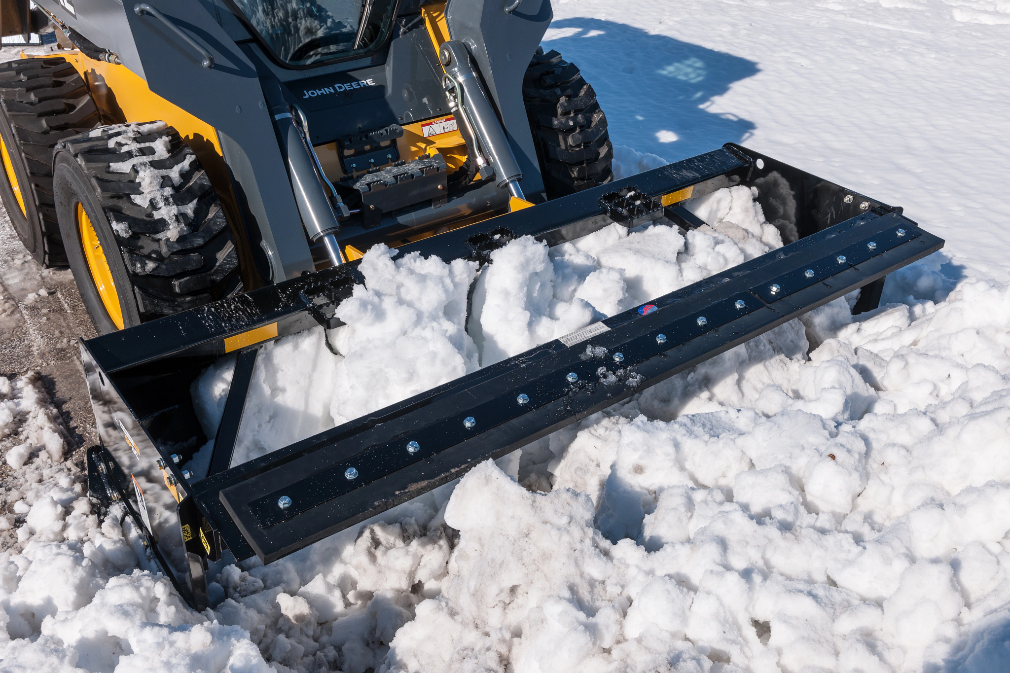 Close-up of skid steer loader using a front-mounted snow pusher to move packed snow, showing the black steel blade filled with snow and the loader’s tires and hydraulic arms.