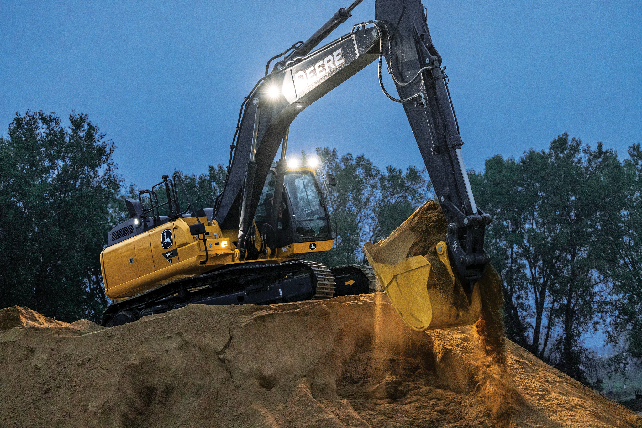 Yellow John Deere excavator with a black arm and illuminated work lights operating at dusk on a sandy dirt mound