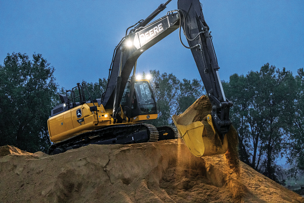 Yellow John Deere excavator with a black arm and illuminated work lights operating at dusk on a sandy dirt mound