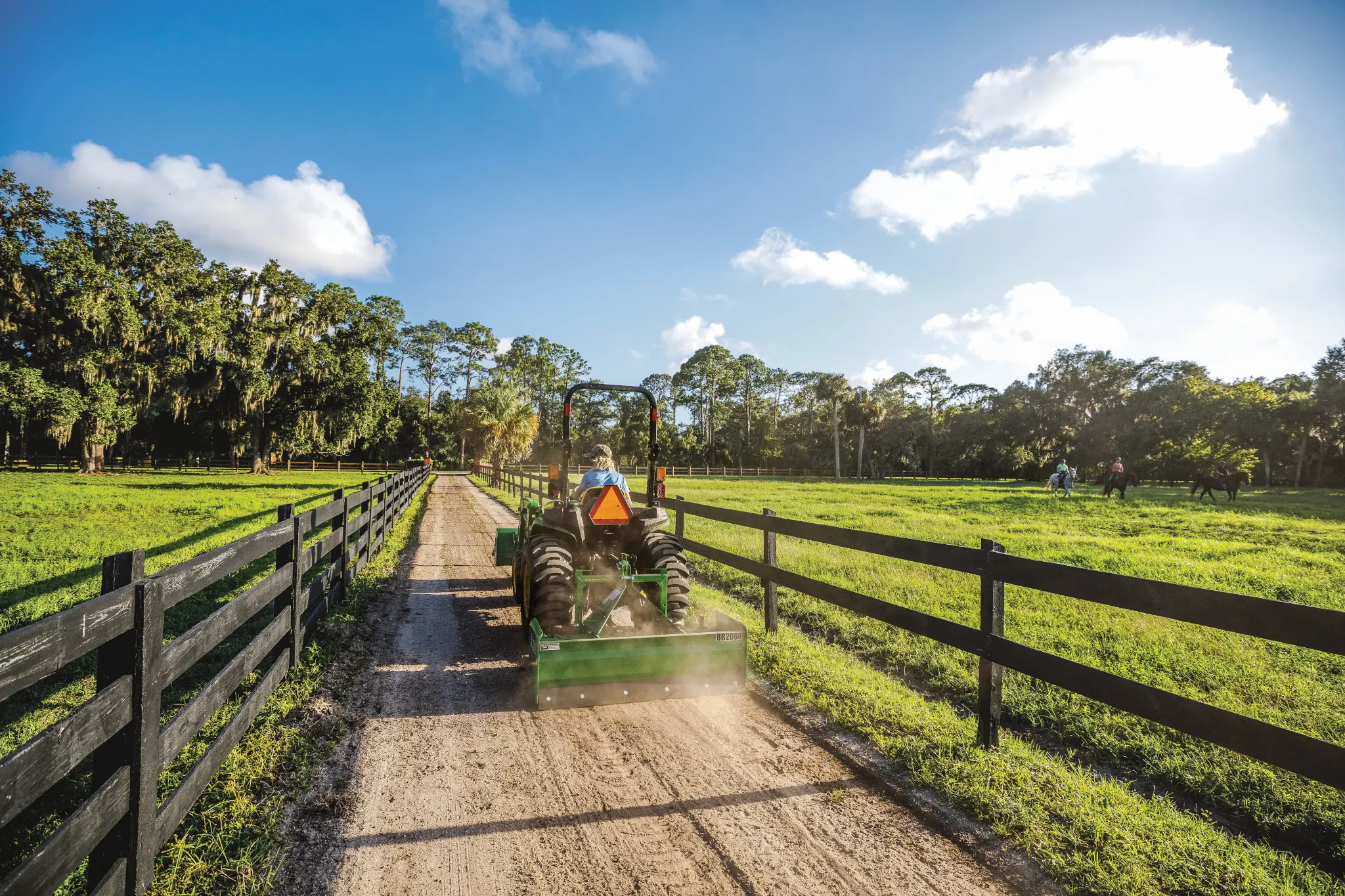 A green tractor levels a dirt lane between wooden fences with a rear land plane, with open grassy fields and trees on both sides under a bright blue sky with scattered clouds