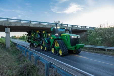 A large tracked tractor towing a folded nutrient applicator down a paved road, passing under a concrete overpass with guardrails on both sides