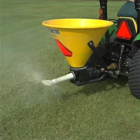 Close-up of a yellow SS1079P pendular spreader at work on green grass. Rear of attached tractor is visible in the right corner.