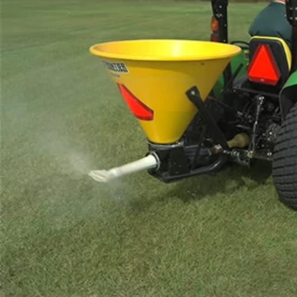 Close-up of a yellow SS1079P pendular spreader at work on green grass. Rear of attached tractor is visible in the right corner.