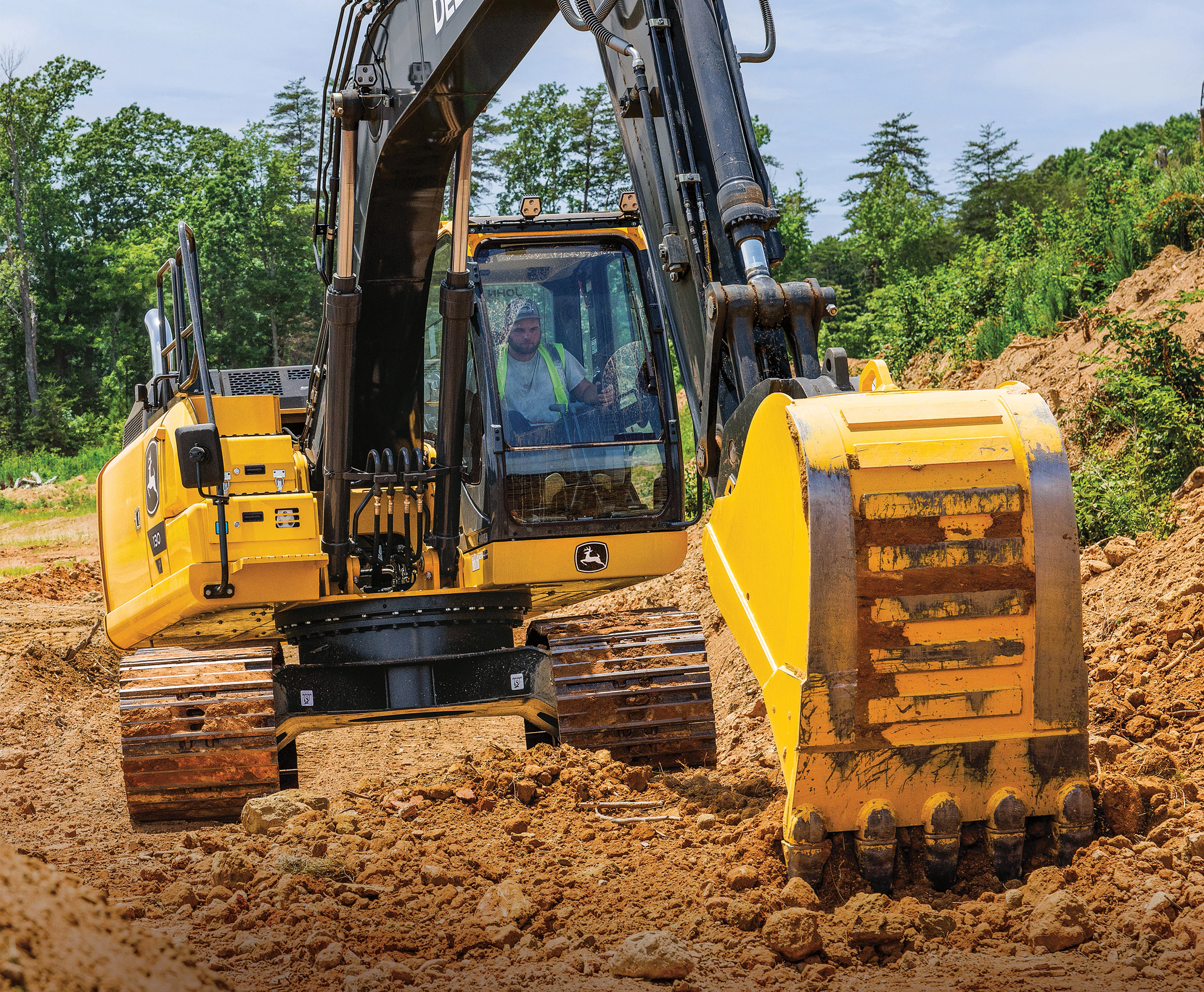 Yellow construction excavator with a large bucket digging into loose brown soil at a worksite, with trees and greenery in the background and an operator visible inside the cab