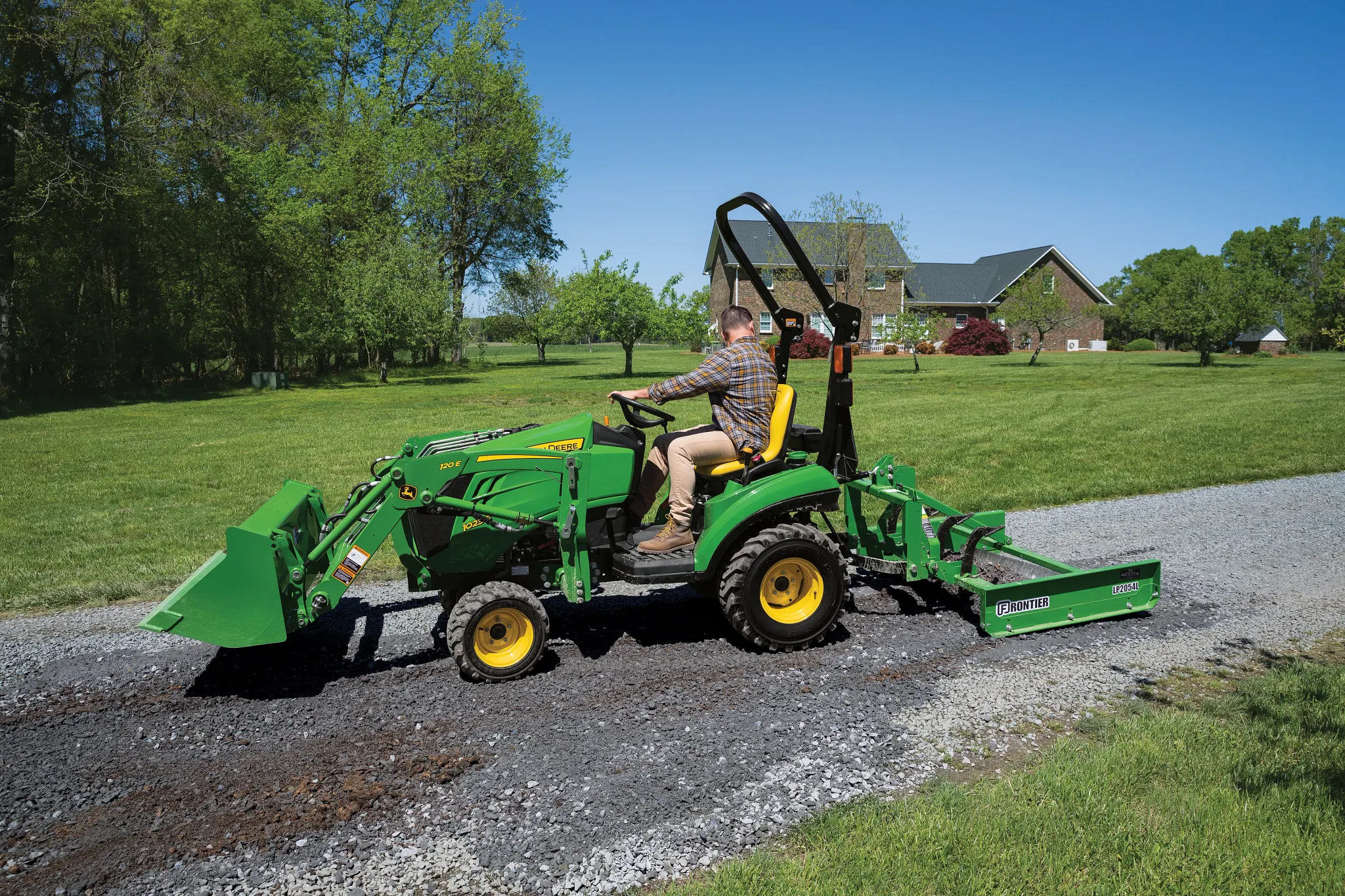 A person drives a small green tractor with a front loader and rear land plane implement along a gravel driveway on a sunny day, with grassy lawn and a brick house in the background