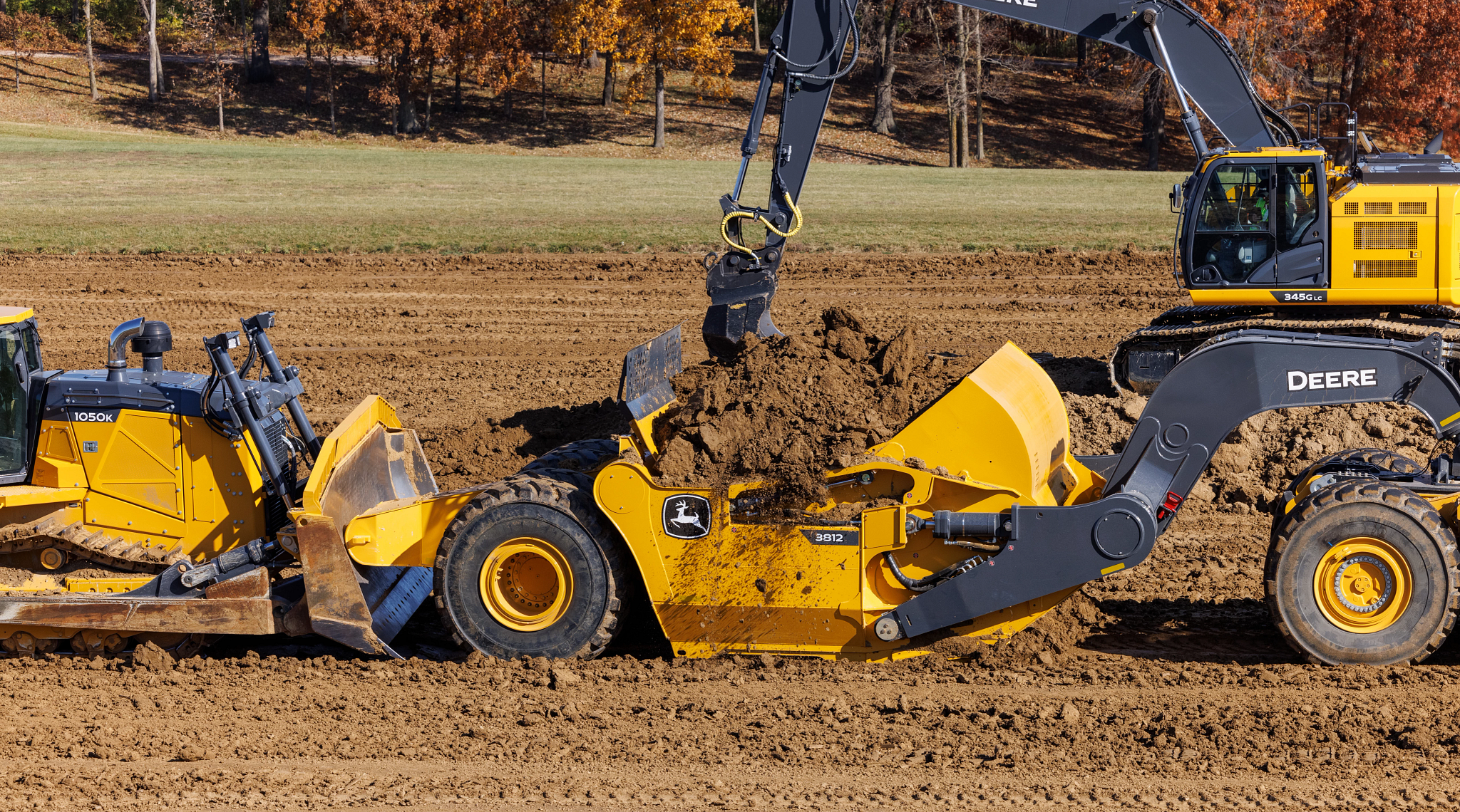 Side view of a 3812 scraper being loaded by a 1050K Dozer and G Series excavator on a dirt and grass jobsite