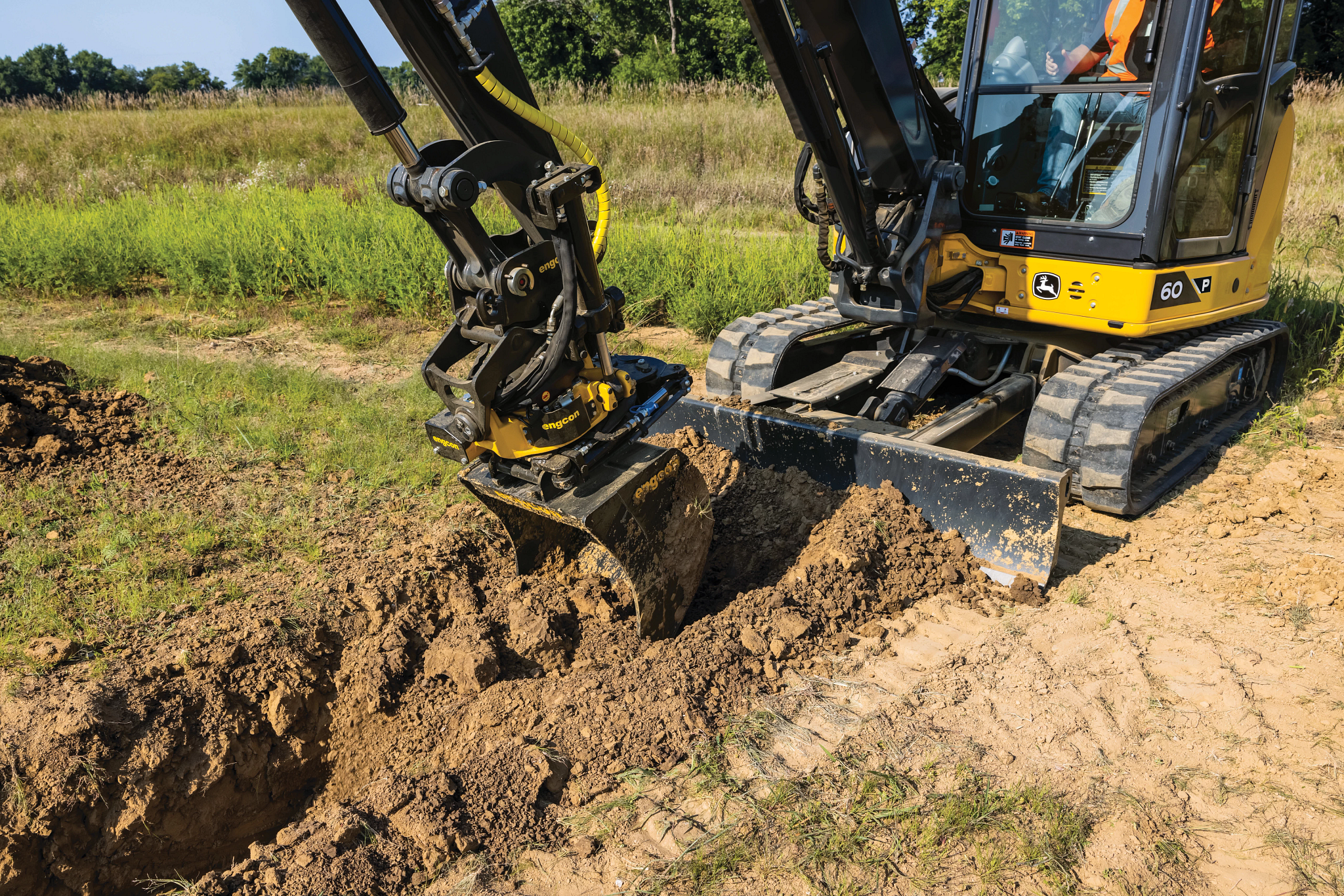 Compact yellow John Deere excavator with a black arm and tilt-rotator attachment digging a narrow trench in sandy soil