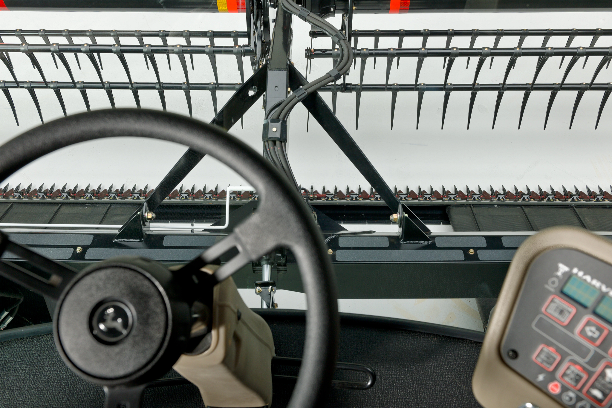 View from inside a windrower cab showing the steering wheel and control panel, looking out toward the header with cutting knives and pickup tines