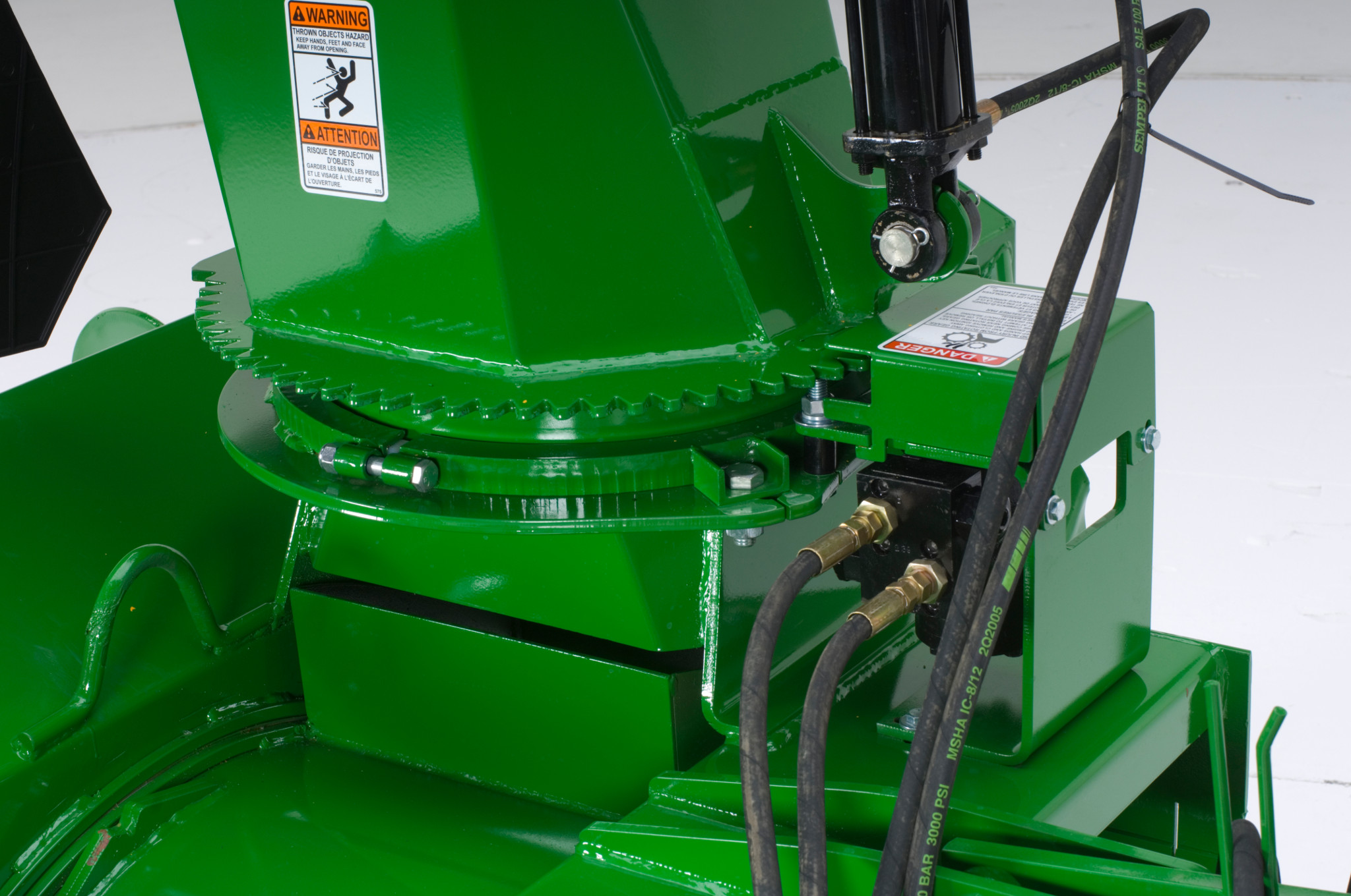 Close-up of a green snowblower housing showing a rotating chute base with gear teeth, hydraulic hoses, and safety decals