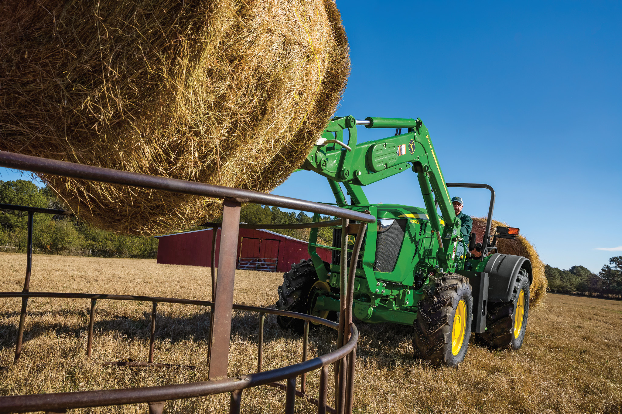 John Deere tractor with front loader lifting a round hay bale over a metal fence in a dry pasture, green body and yellow wheels visible, with farm buildings and open field under a clear blue sky.