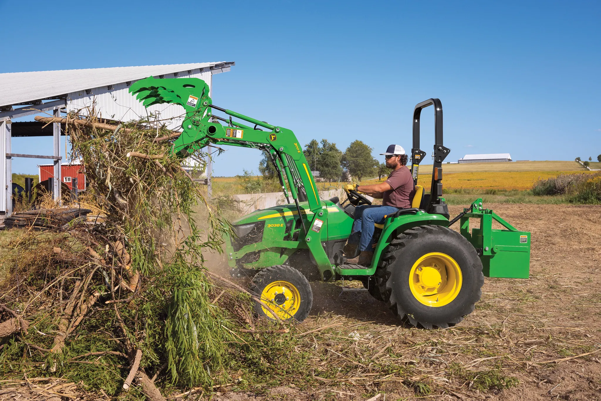 Operator in a green tractor with a front loader and grapple lifts a pile of brush near a metal-sided building, with open farmland stretching into the background under a clear blue sky