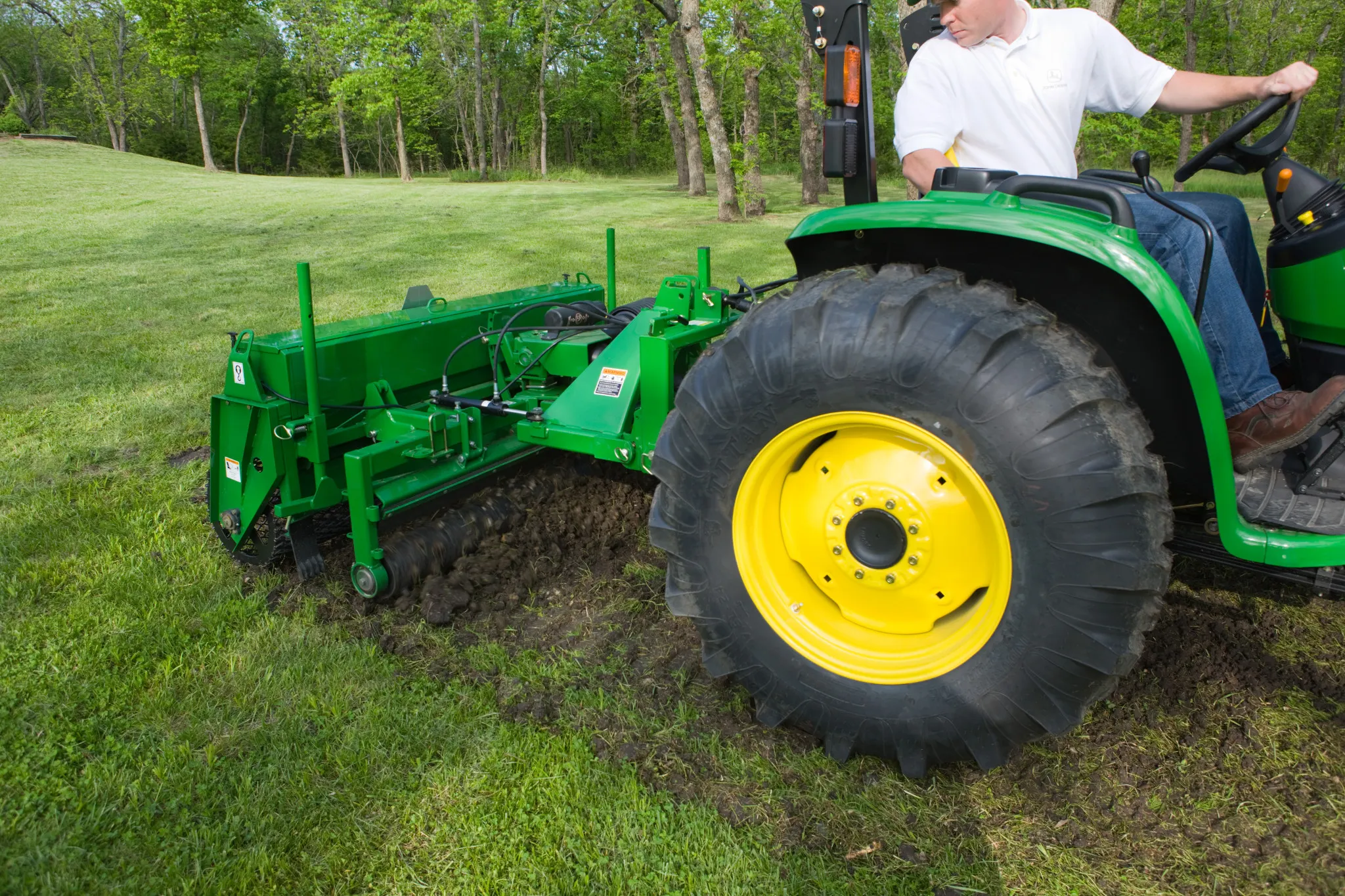 A green tractor with yellow wheels pulls a green soil-cutting attachment that is digging a narrow trench in a grassy field, while a person operates the tractor. Trees and open lawn are visible in the background.