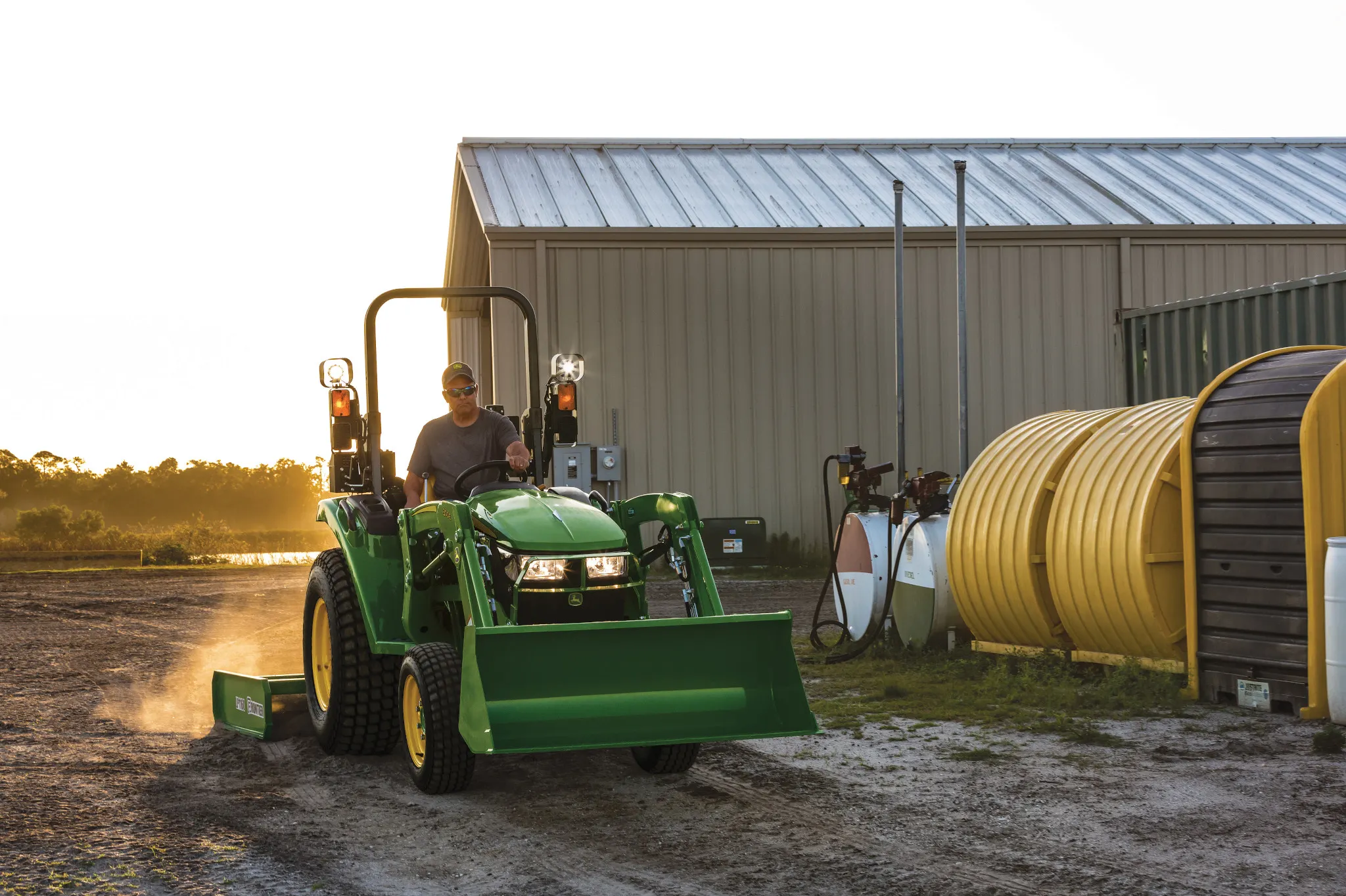 An operator on a green tractor with a front loader and rear land plane drives on a dirt path beside a metal building, with large tanks and equipment nearby at sunset