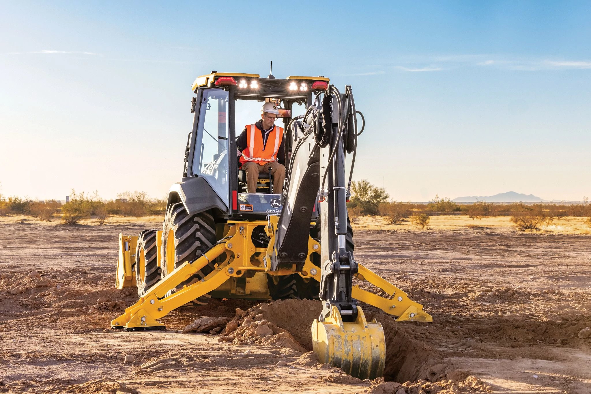 A backhoe loader is positioned over a narrow trench at a flat, open construction site, with stabilizer legs extended on the dirt surface.