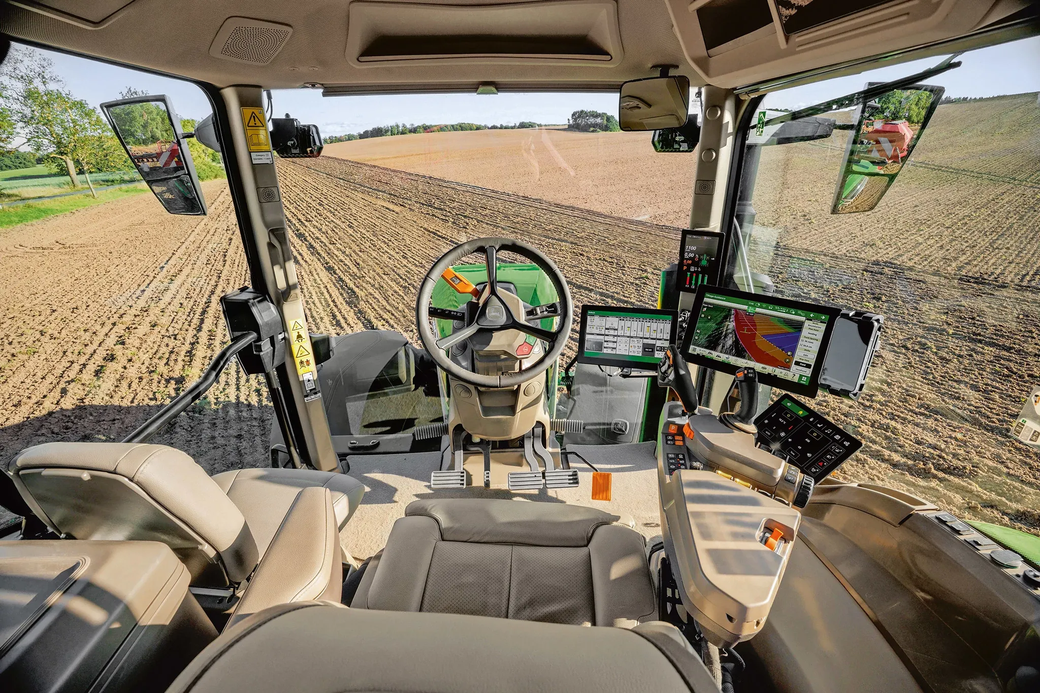 Vue intérieure d’une cabine de tracteur moderne montrant le siège de l’utilisateur, le volant, la console de commande et plusieurs écrans numériques. De grandes fenêtres offrent une vue imprenable sur un champ fraîchement labouré.