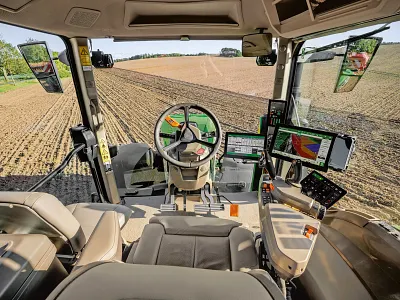 Vue intérieure d’une cabine de tracteur moderne montrant le siège de l’utilisateur, le volant, la console de commande et plusieurs écrans numériques. De grandes fenêtres offrent une vue imprenable sur un champ fraîchement labouré.