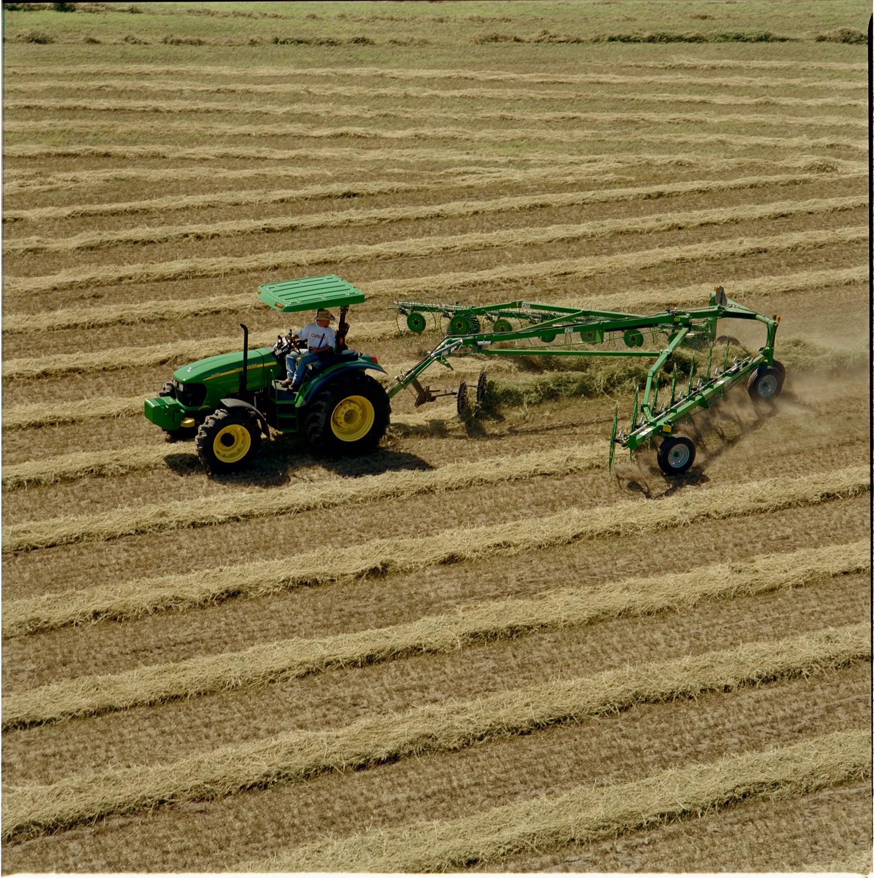 Side left view of a tractor pulling a WR High-Capacity Wheel Rake across a hay field striped with tan windrows