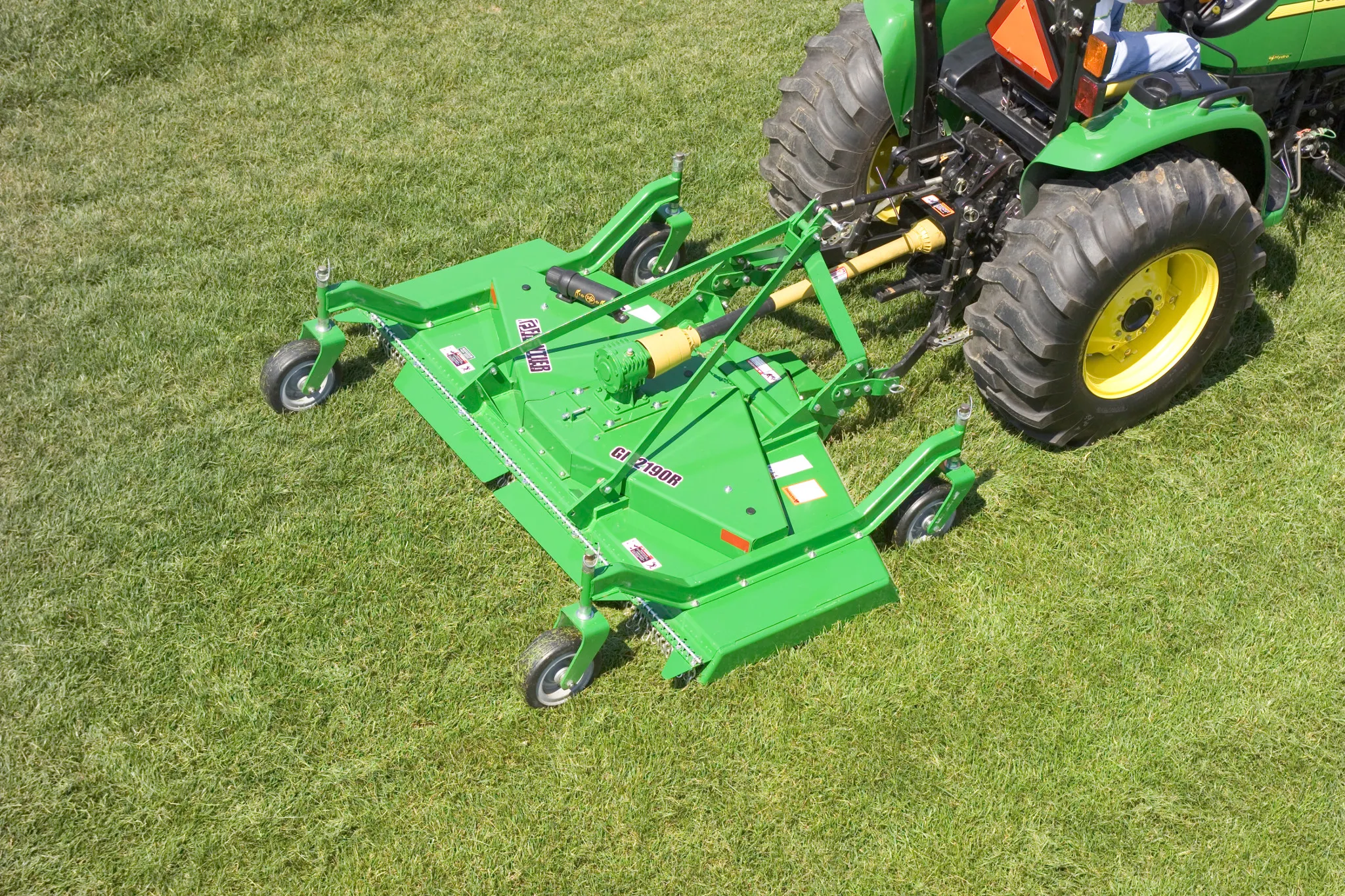 Aerial right view of a green Frontier GM2190R grooming mower on grass with the back end of attached compact tractor in the top right of the image.