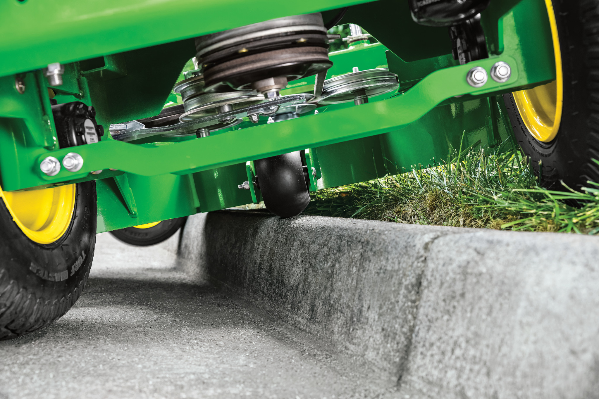 Ground-level close-up of a W36R Commercial Walk-Behind Mower's wheels, green frame, and underside belts as it straddles a curb, with half of the machine on grass and half on pavement