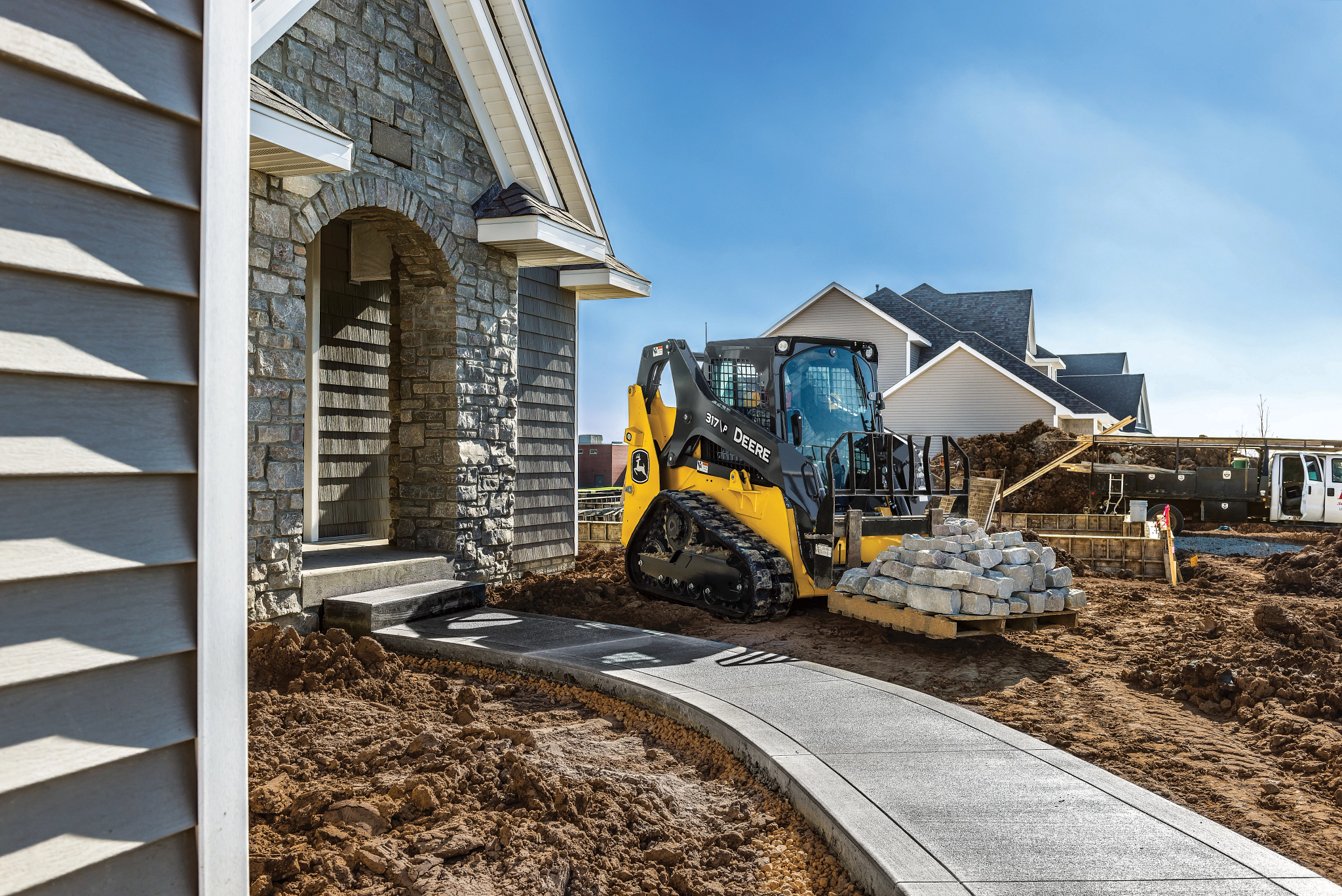 A compact track loader moves a pallet of stone pavers toward the front entrance of a newly built house.