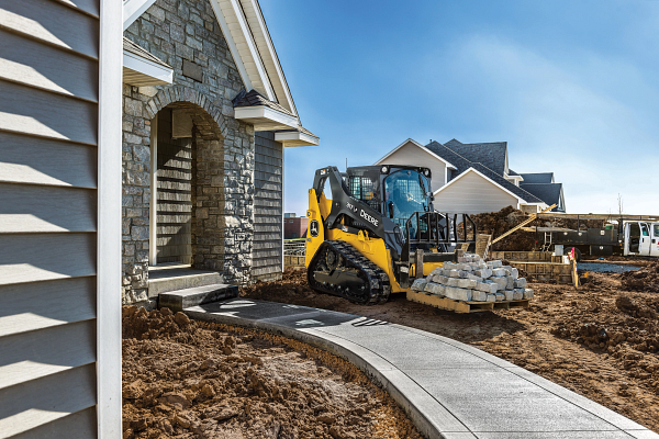 A compact track loader moves a pallet of stone pavers toward the front entrance of a newly built house.