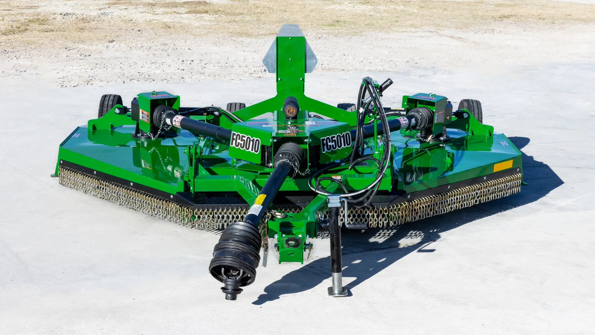 Front view of a green Frontier FC5010 dual-spindle rotary cutter with a PTO driveline, hydraulic hoses, safety chain shielding, and transport wheels, positioned on a concrete surface outdoors