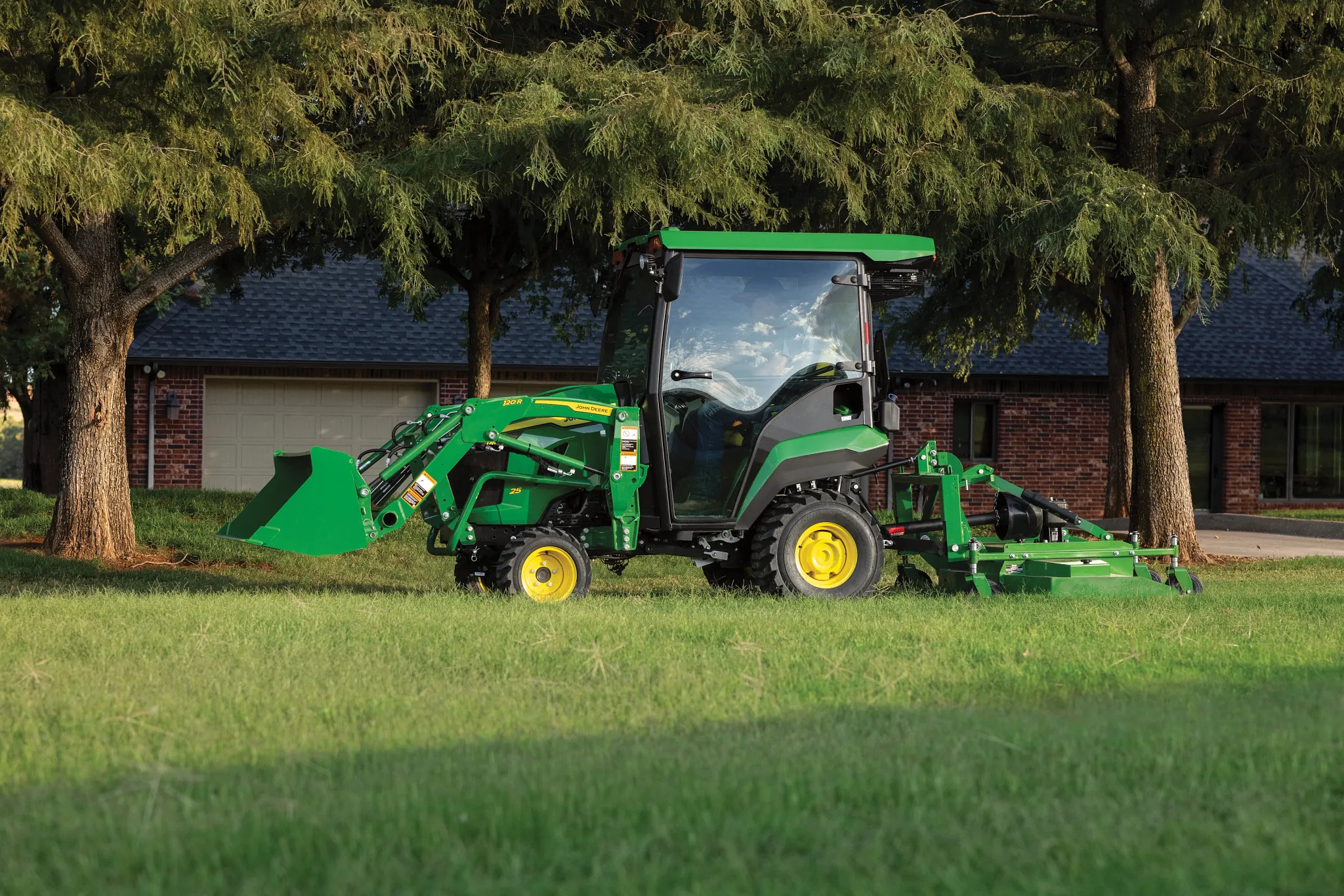 A green tractor with an enclosed cab operates in a grassy yard, using a rear mower attachment to cut grass with a building and large trees in the background