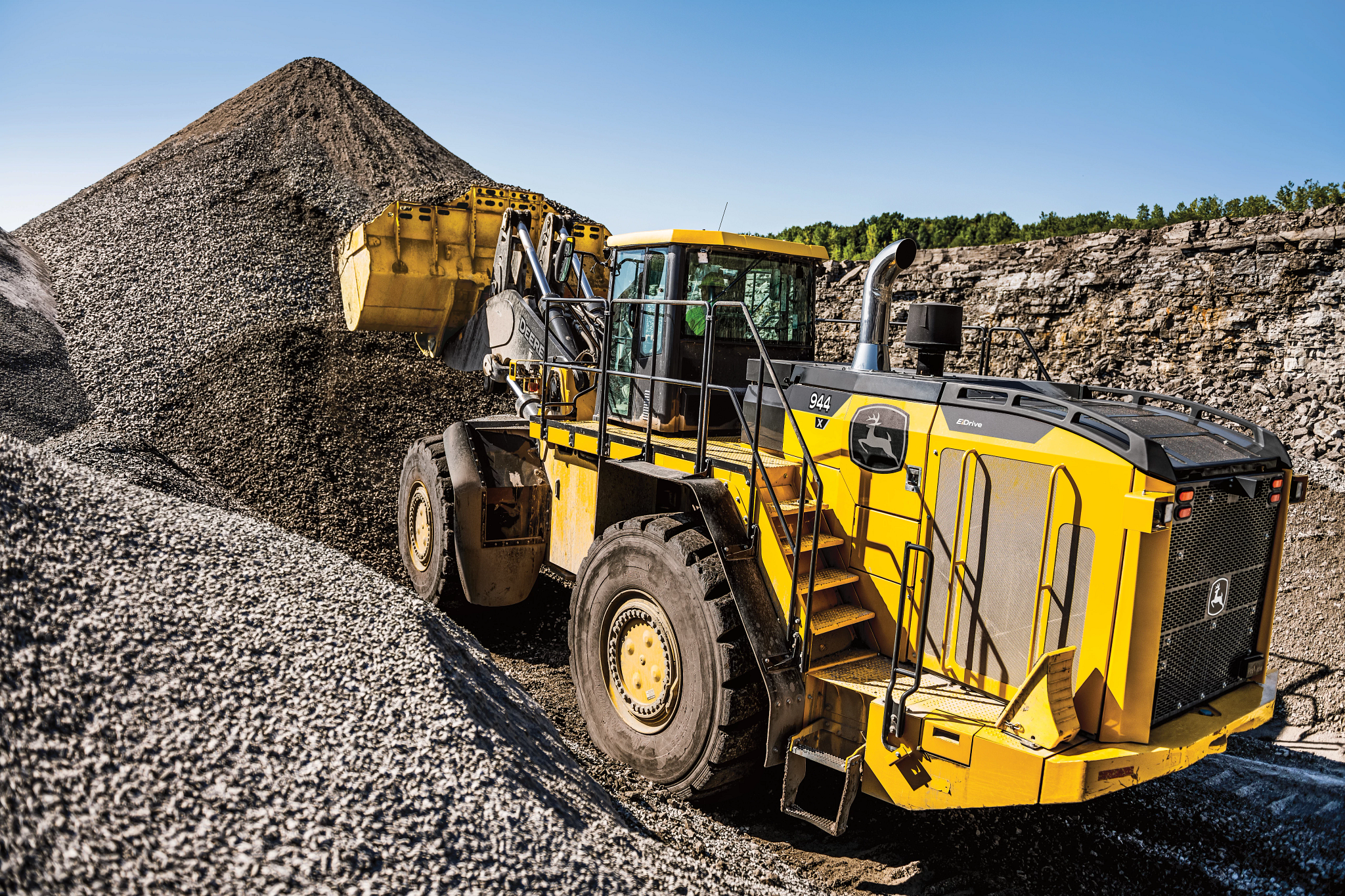 A large yellow wheel loader scoops and carries gravel at a quarry, positioned beside a tall mound of crushed stone