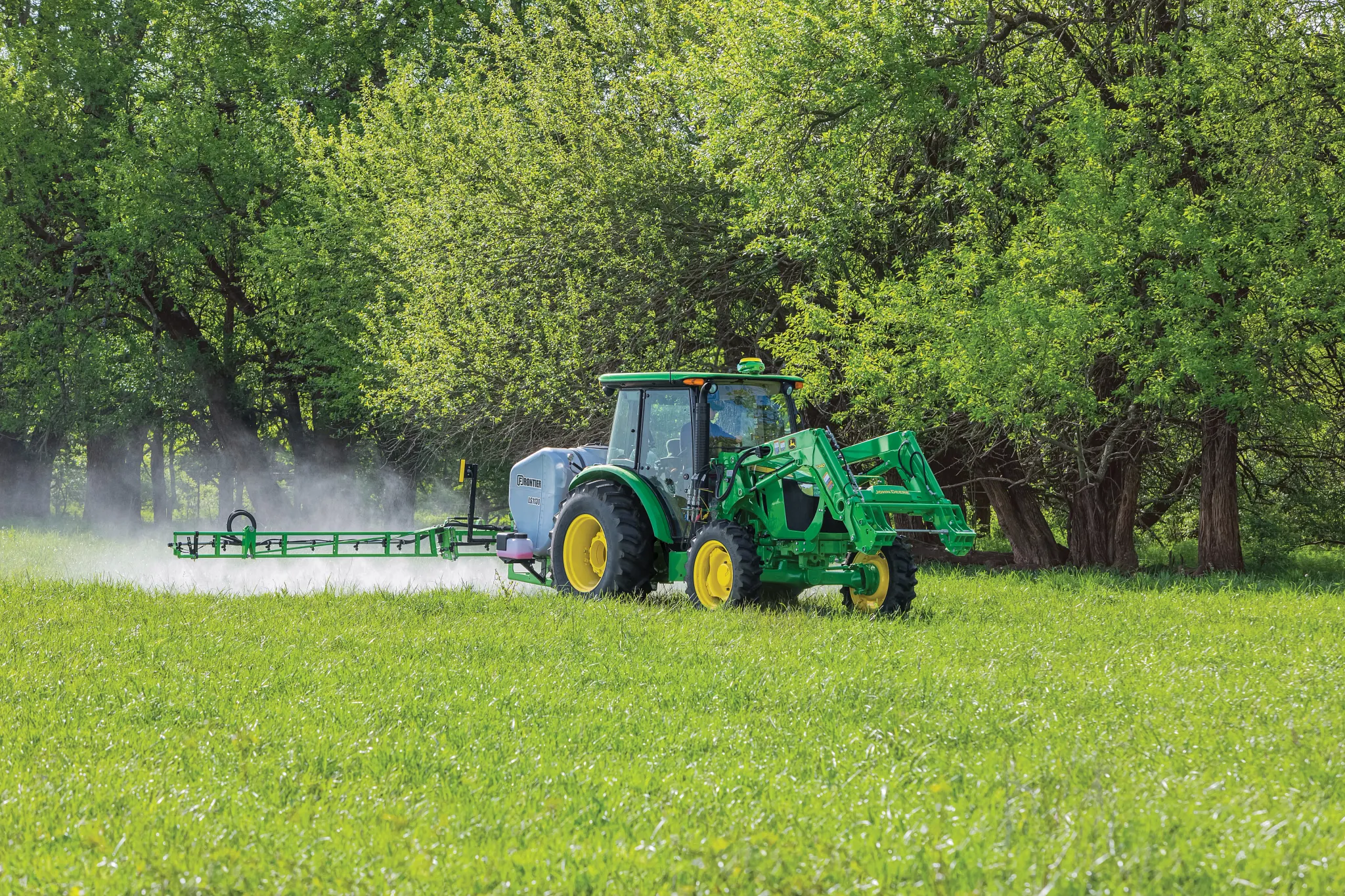 A tractor with a front loader and a wide boom sprayer drives across a grassy field, spraying mist while trees with spring foliage line the background.