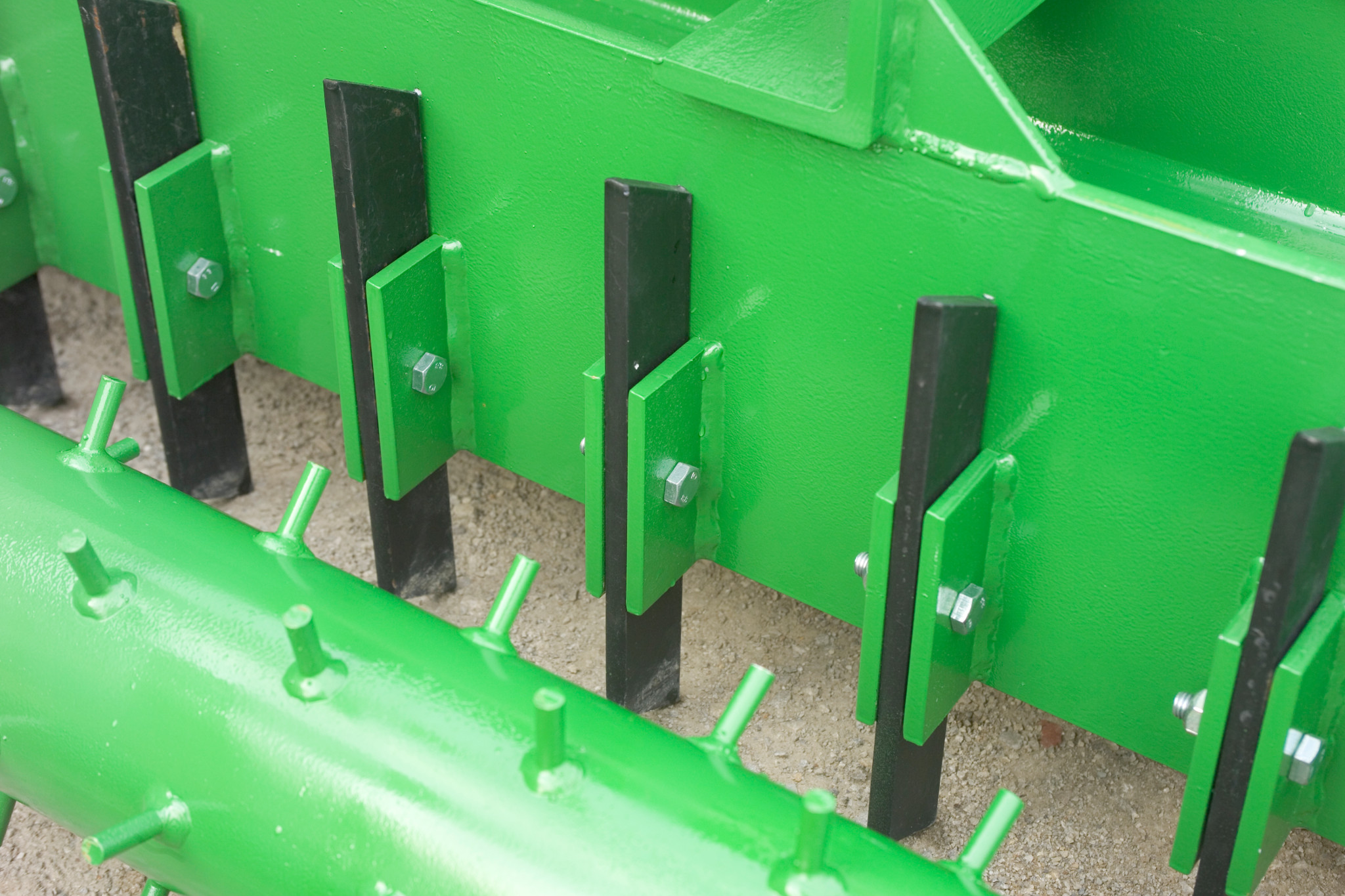 Close-up of green soil pulverizer showing a row of vertical black blades mounted to brackets behind a spiked green roller