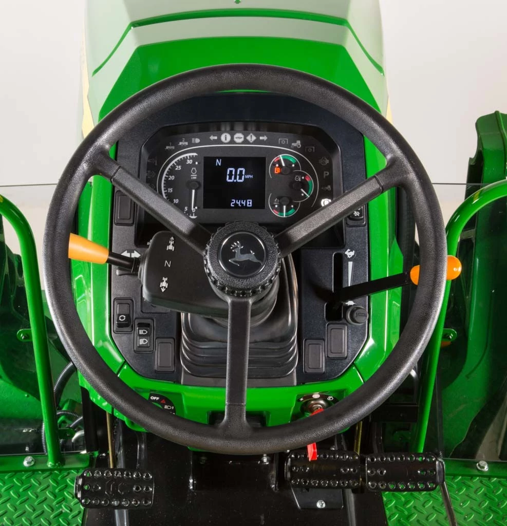 Close‑up operator’s view of John Deere tractor steering wheel with digital instrument cluster, multifunction display showing speed and engine data, turn‑signal stalks, and foot pedals below on a green tractor platform.