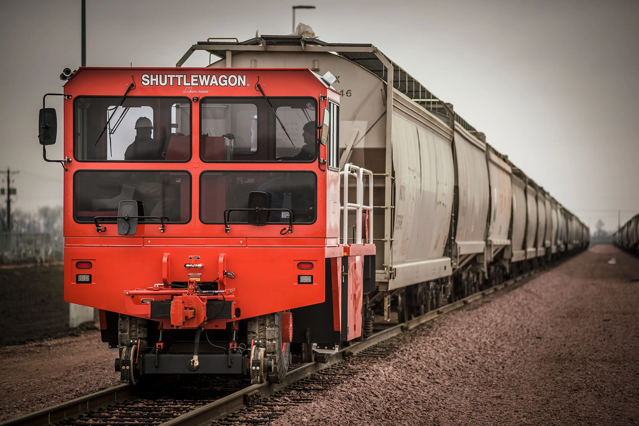 Bright orange rail maintenance vehicle labeled “SHUTTLEWAGON” at the front, coupled to a long line of gray covered hopper freight cars on railroad tracks along a gravel rail bed under an overcast sky
