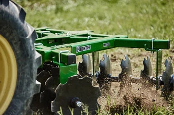 Close-up of a disc harrow being pulled through soil, with angled metal discs cutting into the ground and kicking up dirt in a grassy field