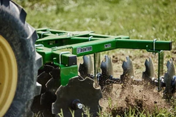 Close-up of a disc harrow being pulled through soil, with angled metal discs cutting into the ground and kicking up dirt in a grassy field