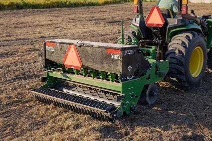 Rear view of tractor pulling a mounted seed drill across a field showing the metal hopper, warning triangle reflectors, and rotating soil-conditioning rollers working the ground.