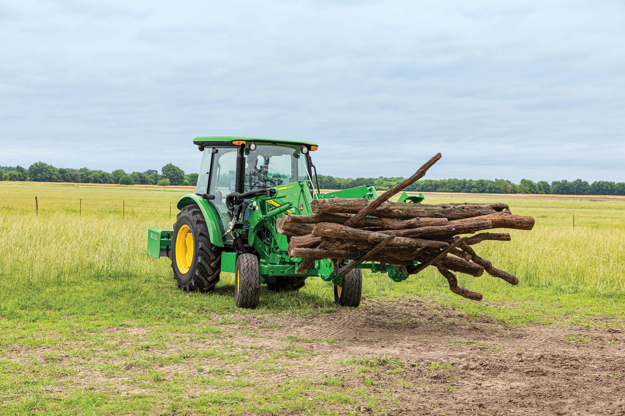 Front right view of a 5075E tractor transporting logs in a grassy field with attached 520M loader and AP12G pallet fork