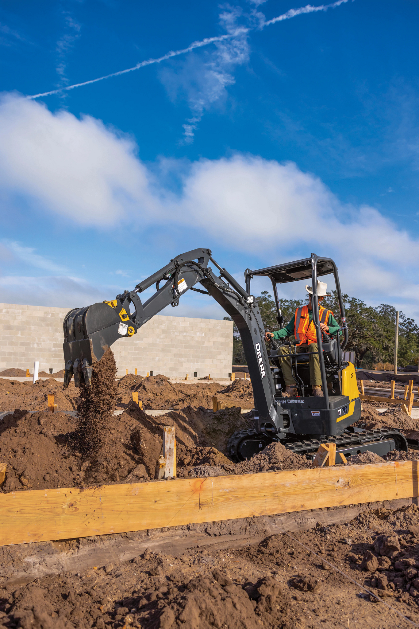 Compact excavator at a construction site dumping soil, with an operator in a high-visibility vest inside the open cab under a bright blue sky