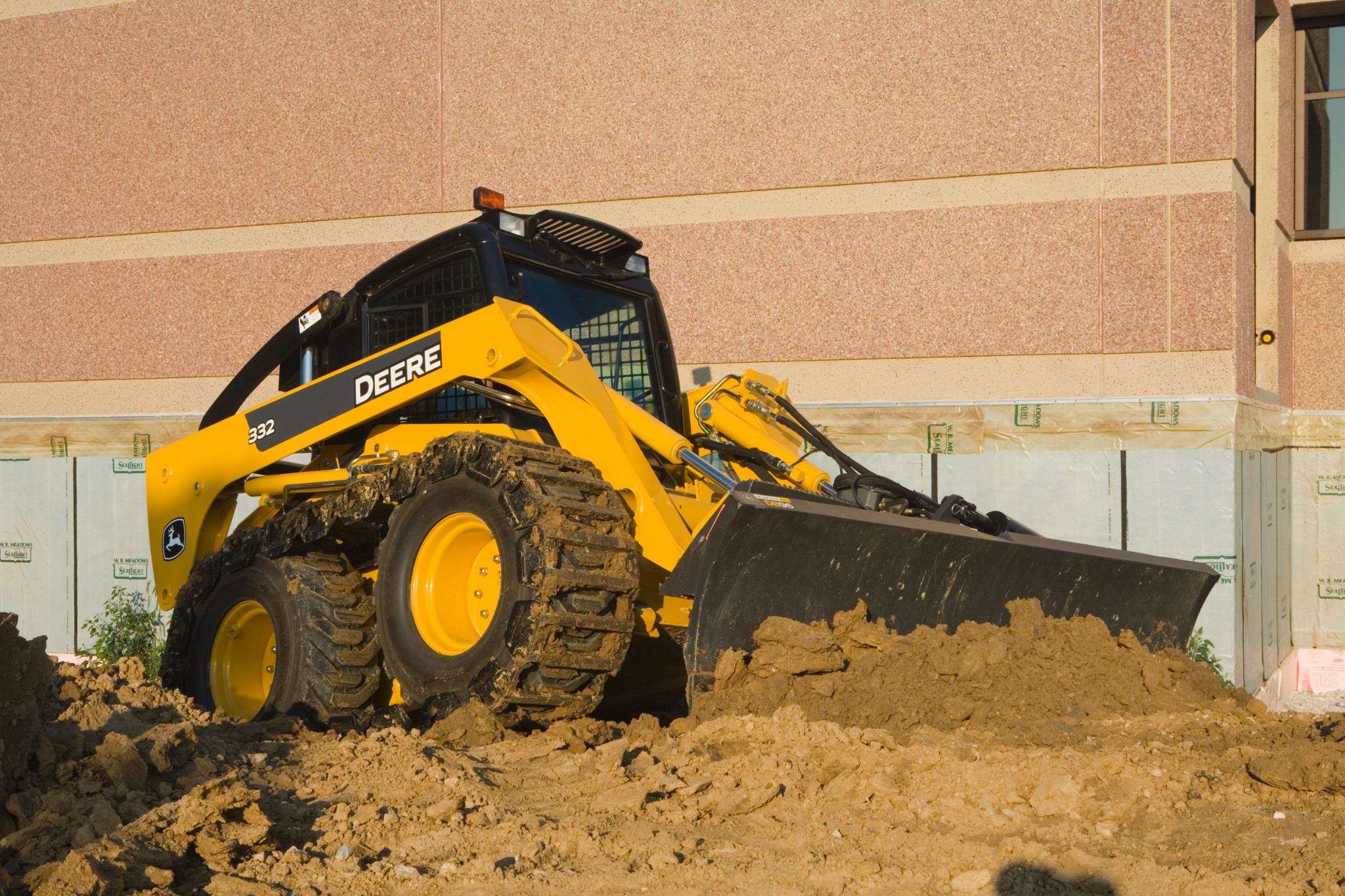 Yellow skid steer loader pushing a large pile of dirt with its front bucket in a construction area beside a tan building