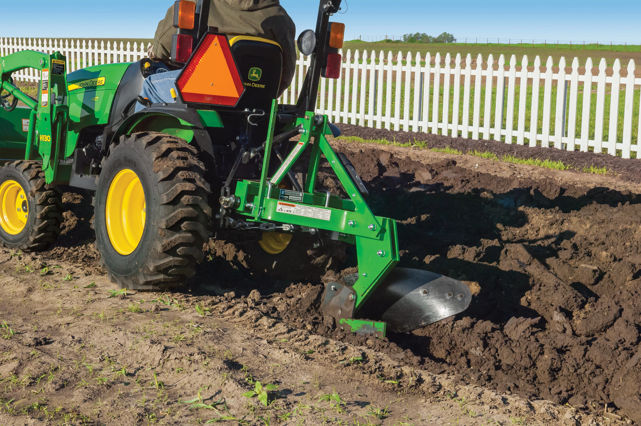 Compact tractor pulling a green turning plow through a garden plot in front of a white picket fence on a sunny day