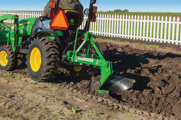 Compact tractor pulling a green turning plow through a garden plot in front of a white picket fence on a sunny day