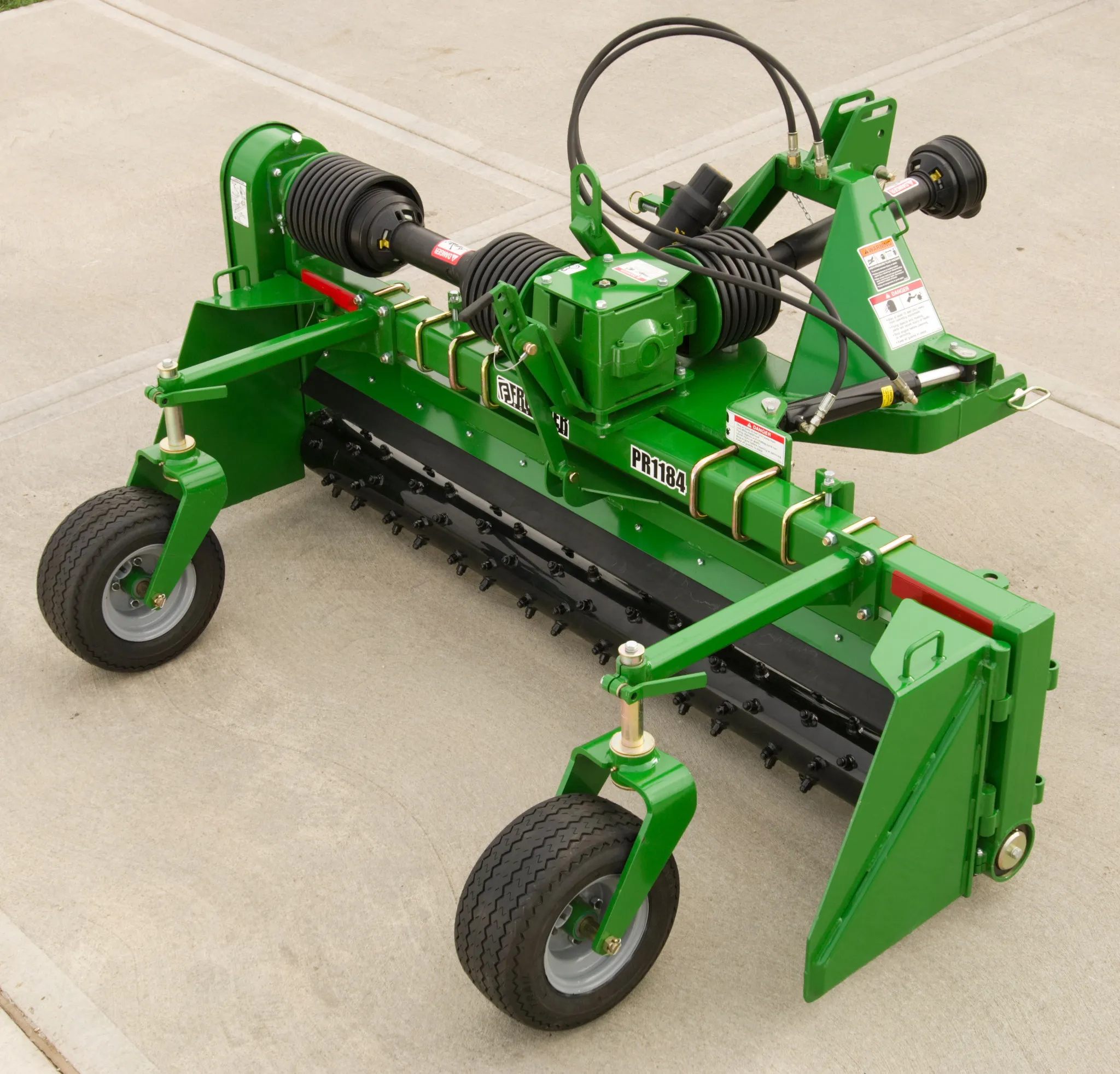 Agricultural mower or brush cutter attachment with a horizontal spinning blade assembly, mounted on two small wheels with adjustable arms, and connected to a power shaft at the top, sitting on a concrete