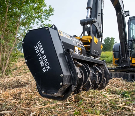 Close-up of forestry mulcher attachment mounted on an excavator arm, with rotating cutting drums and visible safety warning text reading “KEEP BACK 300 FT / 90 M”.