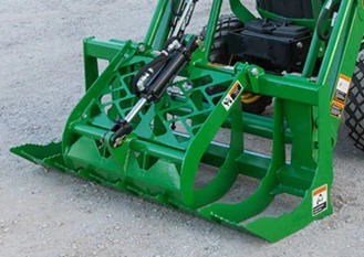 Front left view of a green root grapple with claws closed resting on gravel with the front of its attached tractor in the back corner