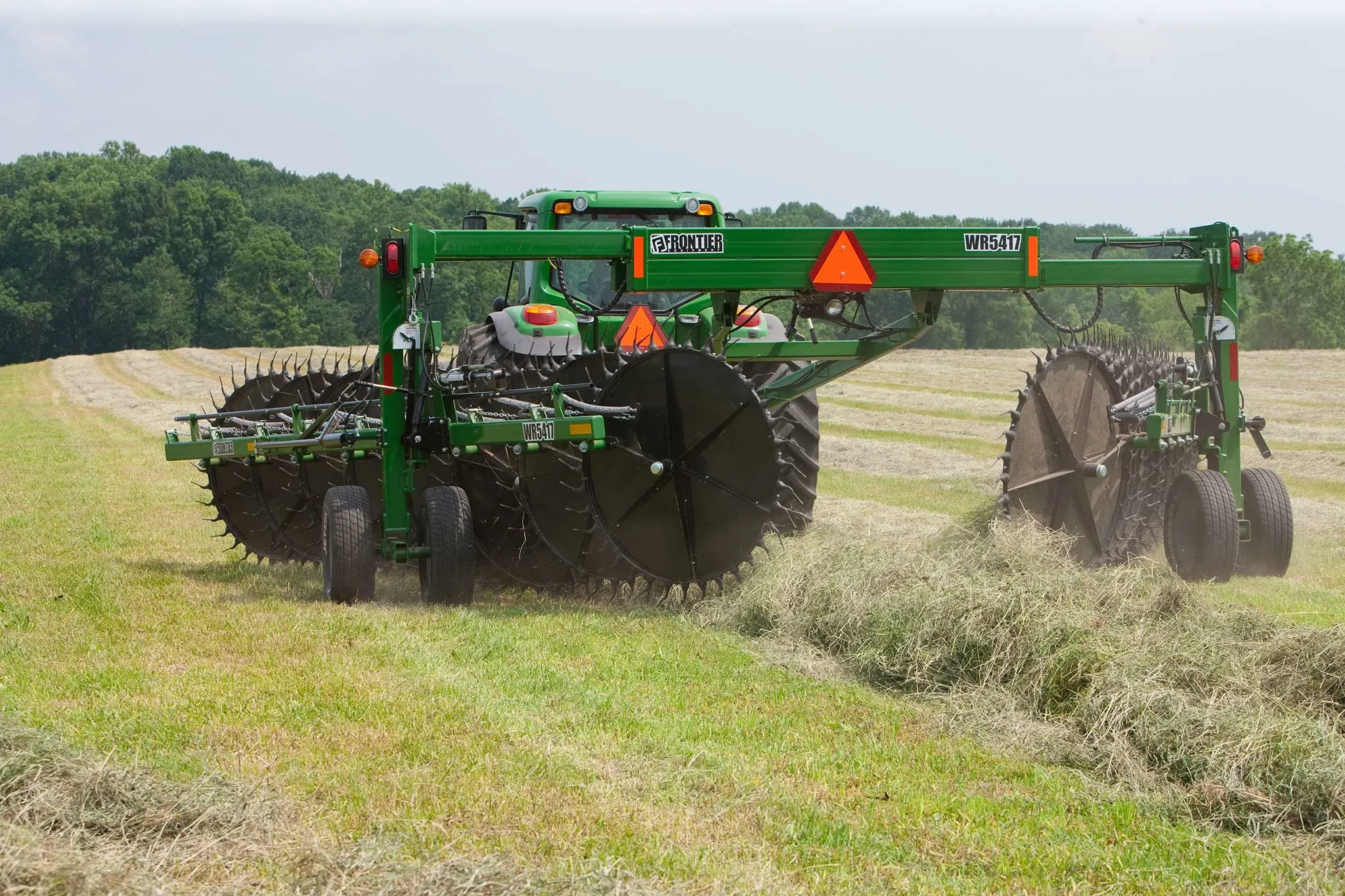 Rear view of a WR5417 wheel rake and tractor raking windrows in a tree-lined hay field