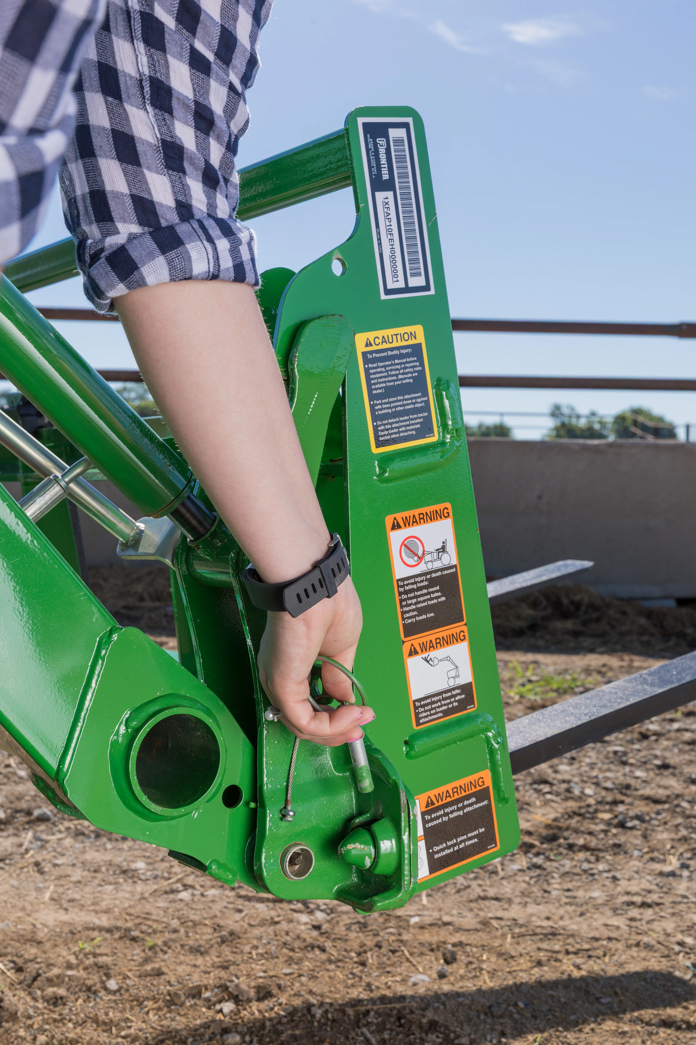 Close-up of a a safety locking pin with cable being inserted into a green tractor or loader attachment bracket, showing warning labels, hydraulic linkage, and metal mounting points.