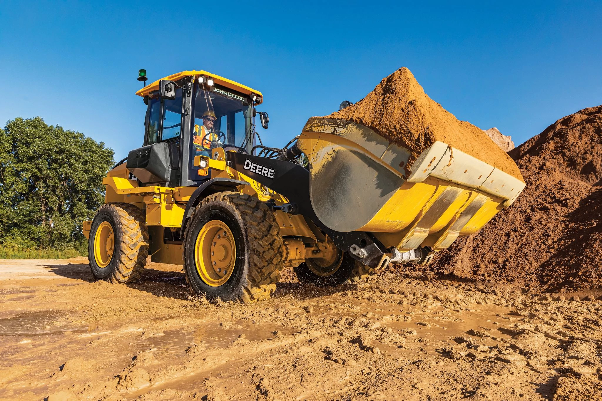A front-end loader sits on a dirt construction site under a clear blue sky, holding a full bucket of soil raised slightly off the ground.