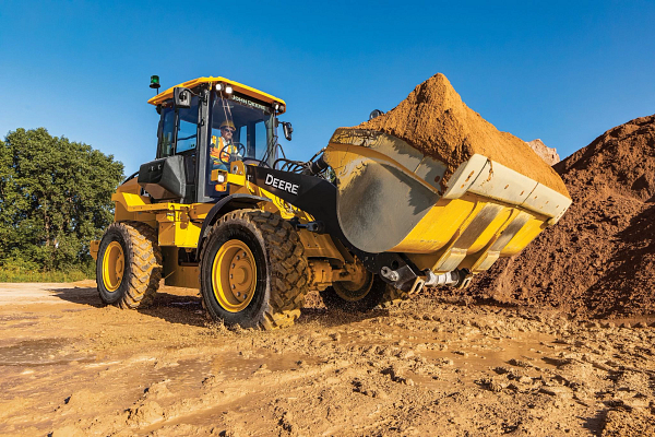 A front-end loader sits on a dirt construction site under a clear blue sky, holding a full bucket of soil raised slightly off the ground.